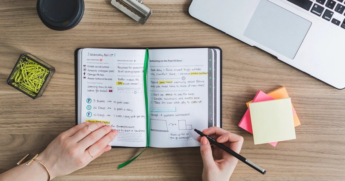 Hands writing in a notebook full of goals and notes, with a laptop beside it: anyone starting online trading learns more by writing down their own rules and keeping a trading journal